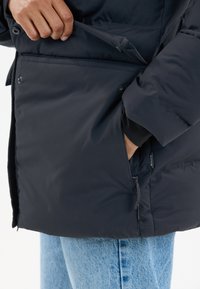 Person trägt eine schwarze Steppjacke, mit einer Hand in einer seitlichen Reißverschlusstasche und der anderen Hand, die eine Druckknopfklappe anpasst.