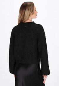 Maglione nero corto con texture a coste, maniche ampie e scollo a girocollo. Il design a maglia ha un leggero luccichio, abbinato a una gonna nera in satin.