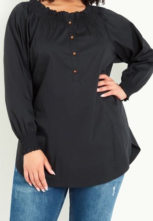 Kobieta w czarnej bluzce z długim rękawem, ozdobionej drewnianymi guzikami i marszczonymi mankietami, w połączeniu z postrzępionymi niebieskimi dżinsami, z ręką na biodrze.