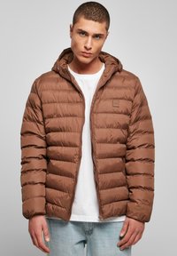 Bruine pufferjack met een ritsluiting, capuchon en horizontaal gestikte patroon. G gedragen over een wit T-shirt met lichtblauwe jeans.