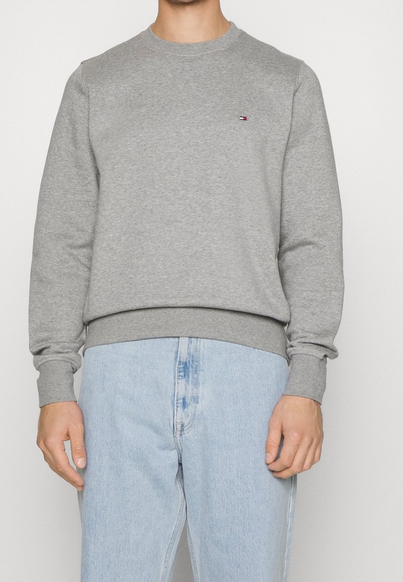 Homme portant un sweat-shirt gris uni à manches longues avec un petit logo Tommy Hilfiger sur la poitrine et un jean bleu clair.
