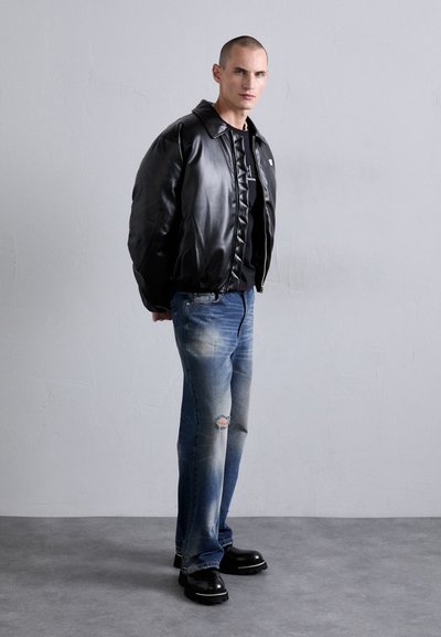 Veste bomber en cuir noir avec fermeture éclair, associée à un jean en denim bleu délavé comportant des détails usés et des chaussures noires imposantes.