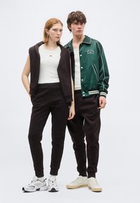 Νεαρός άνδρας και γυναίκα που φορούν ταιριαστά μαύρα παντελόνια jogging και λευκά μπλουζάκια, στέκονται δίπλα-δίπλα, η γυναίκα με μαύρο φούτερ με κουκούλα κρεμασμένο στους ώμους.