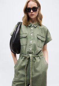 Γυναίκα που φοράει πράσινη ολόσωμη φόρμα με κουμπιά και ζώνη, μαύρα μεγάλα γυαλιά ηλίου, χρυσούς κρίκους στα αυτιά και κρατά μια μεγάλη μαύρη τσάντα ώμου.