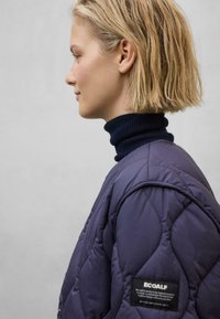Lilla quilted jakke med højhalset marineblå turtleneck. Har et logo-patch på ærmet og blødt, tekstureret stof.