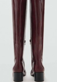 Burgundin yli polven saappaat, jotka on valmistettu sileästä nahasta kiiltävällä pinnalla. Saappaissa on matala lohko korko ja sivuvetoketjut helppoa käyttöä varten.