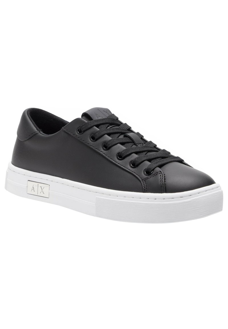 Black leather sneaker with round toe, flat white rubber sole, and black laces. Features a logo detail on the side and smooth texture.