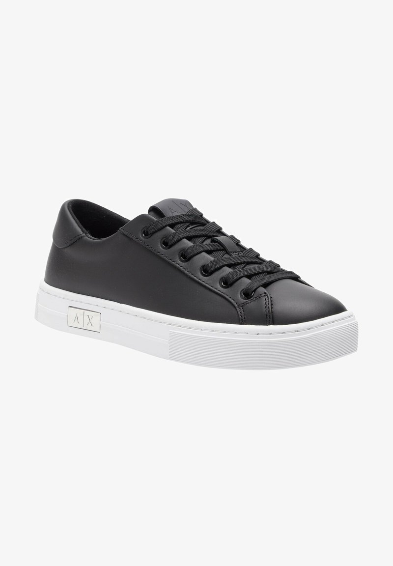 Black leather sneaker with round toe, flat white rubber sole, and black laces. Features a logo detail on the side and smooth texture.
