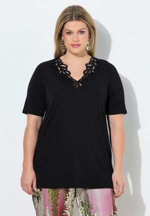 Kobieta ubrana w czarny krótki top z wycięciami w kwiatowy wzór na dekolcie oraz w wielokolorowe spodnie w kwiaty na białym tle.