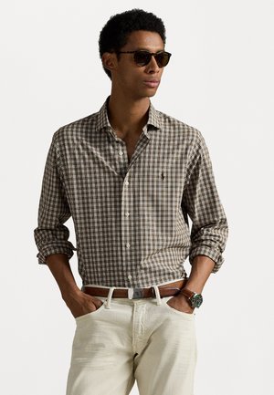 Braun karierte Button-Up-Hemd mit hochgekrempelten Ärmeln, kombiniert mit cremefarbenen Hosen und einem braunen Gürtel. Metallarmbanduhr und dunkle Sonnenbrille.