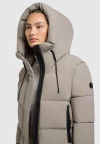 Veste matelassée isolante beige avec une grande capuche, fermeture éclair noire et quincaillerie assortie, ainsi que des poches latérales. Elle présente un design matelassé et des cordons de serrage ajustables.