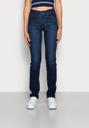 Dark blue denim jeans with a slim fit and minimal stitching details, paired with white sneakers on a plain background.
