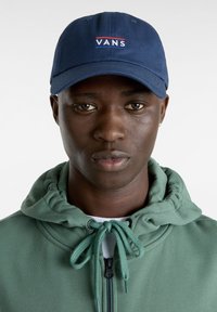 Navy blue baseballcap med "VANS" logo i hvid og rød, lavet af bomuld. Båret med en grøn zip-up hoodie med snøre.
