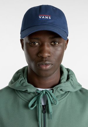 Casquette de baseball bleu marine avec le logo "VANS" en blanc et rouge, fabriquée en coton. Portée avec un sweat à capuche vert zippé avec cordon de serrage.