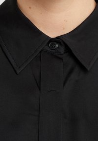Zwart collared shirt met een gladde textuur, een enkele knoopsluiting en subtiele stiksels langs de kraag en de knooplijst.