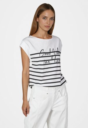 Young woman wearing a white sleeveless top with black horizontal stripes and text, paired with white pants, standing against a plain background.