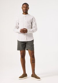 Camisa de rayas blanca de manga larga con cuello de banda, combinada con pantalones cortos de color verde olivo. Zapatos de ante marrón tipo slip-on y tatuajes visibles en las pantorrillas.