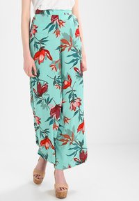 Kwiatowe spodnie o szerokiej nogawce w miętowozielonym materiale z czerwonym i zielonym wzorem botanicznym. Posiadają wysoki stan i zwiewny krój. Noszone z beżowymi butami na platformie.