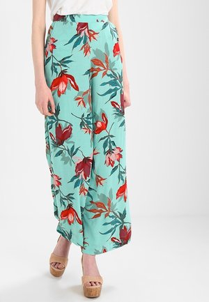 Pantalons larges fleuris en tissu vert menthe avec imprimé botanique rouge et vert. Caractéristiques : taille haute et design fluide. Portés avec des chaussures à plateforme beiges.