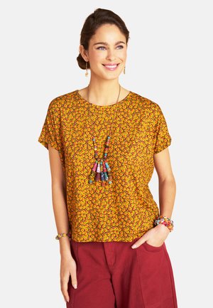 Frau lächelt, trägt ein gelbes, gemustertes kurzärmeliges Oberteil, bordeauxrote Hose, eine bunte Perlenkette, Ohrringe und Armbänder.