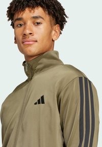 Olivgrön dragkedjans jacka gjord av slät tyg, med svarta tre-randiga detaljer på axlarna och en liten svart logotyp på bröstet.