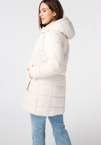 Pufferjas in lichtbeige met capuchon, voorzien van een doorgestikt ontwerp, zijzakken en een ritssluiting. De stof lijkt zacht en warm te zijn.