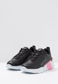 Baskets de sport noires avec une surface texturée, semelle mouchetée blanche et une accentuation rose sur le talon. Présentent un design à lacets et une languette rembourrée.