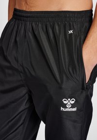 Zwarte sportbroek gemaakt van glanzende, lichtgewicht stof. Voorzien van een elastische tailleband, zijzakken en een wit logo aan de voorkant.
