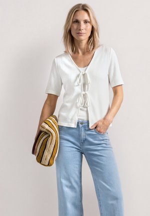 Mujer con top blanco de manga corta con detalles de lazo en el frente y vaqueros azul claro, sostiene un jersey de rayas y está de pie frente a un fondo liso.