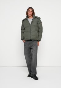 Green puffer jacket with a zip front, large collar, and side pockets. Paired with grey jeans and black footwear, against a plain background.