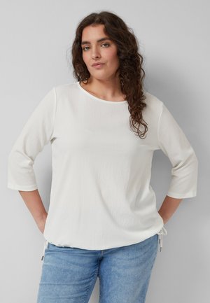 Kobieta o kręconych brązowych włosach, ubrana w biały top z rękawami do łokcia i niebieskie dżinsy na jasnoszarym tle.