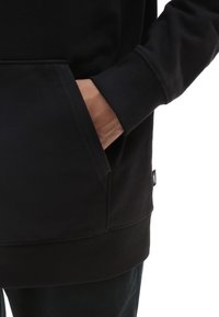 Mano parzialmente nella tasca anteriore di una felpa nera a maniche lunghe con polsini a costine e un piccolo etichetta di stoffa del marchio.