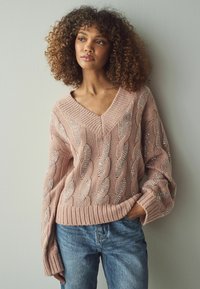 Hellrosa Kabelstrickpullover mit V-Ausschnitt und silbernen Verzierungen, kombiniert mit blauen Jeans. Strukturiert, lockere Passform mit gerippten Bündchen.