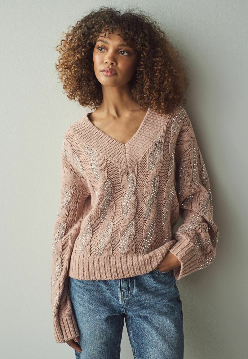 Hellrosa Kabelstrickpullover mit V-Ausschnitt und silbernen Verzierungen, kombiniert mit blauen Jeans. Strukturiert, lockere Passform mit gerippten Bündchen.