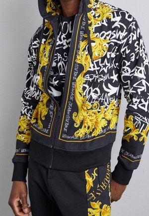 Schwarzer Zip-Hoodie und Hose mit gelben Barockmustern und weißem Graffiti-Stil-Text, getragen von einer Person vor einem schlichten grauen Hintergrund.