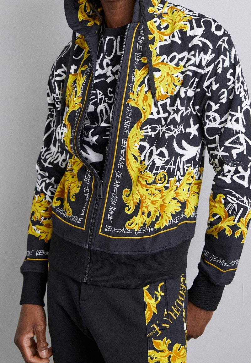 Sweat à capuche noir zippé et pantalon avec des motifs baroques jaunes et un texte de style graffiti blanc, portés par une personne sur un fond gris uni.