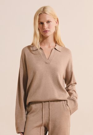 Donna bionda che indossa un top beige a maniche lunghe in maglia con colletto e pantaloni beige in maglia abbinati con coulisse in vita.