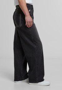 Jeans de mezclilla negros de pierna ancha con detalles de costura blanca, que cuentan con bolsillos traseros y una textura suave. Combinados con zapatillas blancas.