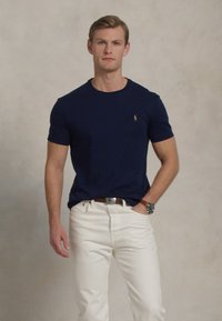 Man som bär en marinblå kortärmad skjorta med liten logotyp, vita byxor, brun bälte och en armbandsur, stående mot en neutral bakgrund.