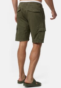 Hombre vistiendo pantalones cortos de cargo verde oliva y sandalias a juego, de pie con la espalda hacia la cámara sobre un fondo blanco.