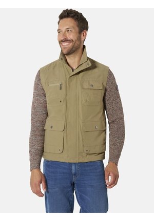 Man wearing beige utility vest over brown knitted sweater and blue jeans, smiling and looking to the side against white background.