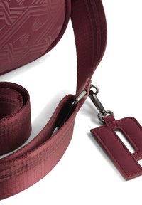 Burgundy axelväska med geometrisk textur, tygrem, metallbeslag och en tagg med en utstansad bokstav "P."