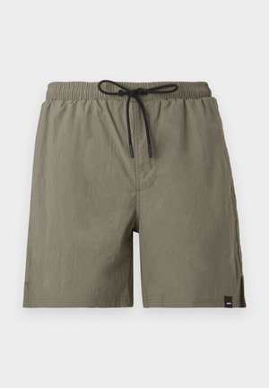Homme debout portant une chemise sombre à manches courtes avec boutons, un short gris clair et des chaussures bateau en cuir marron sur un fond uni.