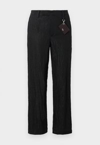 Černé, strukturované kalhoty s širokým střihem a hladkým povrchem, viditelné švy; obsahují štítek připevněný stříbrným kovovým kroužkem.