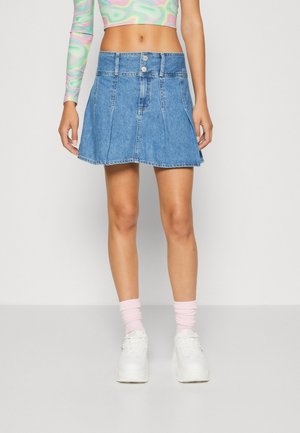 Kvinde iført en blå plisseret denim mini nederdel, pastel swirl langærmet cropped top, pinke sokker og hvide sneakers, stående mod hvid baggrund.