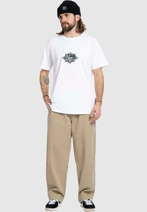 Hvit bomulls T-skjorte med svart geometrisk grafikk i midten, kombinert med løse beige bukser og svart lue. Svarte og hvite sneakers.