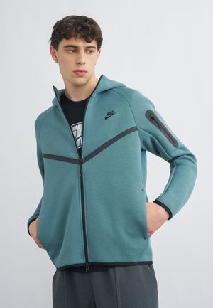 Mladý muž v tyrkysovej mikine na zips Nike s čiernymi akcentmi, ruky v vreckách, pozerá sa doľava na jednoduchom svetlom pozadí.