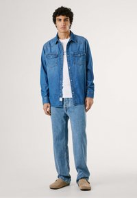 Denimskjorta i medelblått med två bröstfickor, knäppning med knappar och kontrasterande sömmar. Kombinerad med ljusblå jeans och tan färgade skor.