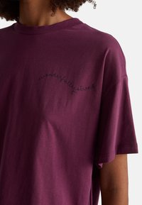 Burgundowy t-shirt z bawełny z okrągłym dekoltem i krótkimi rękawami, ozdobiony czarnym, kursywnym haftem, który głosi "cudownie silny".