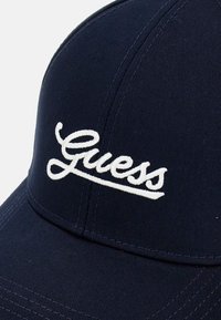 Mørkeblå baseballkasket lavet af tekstureret stof, der har et hvidt broderet "Guess" logo på fronten. Kurvet skygge og syet detaljer.